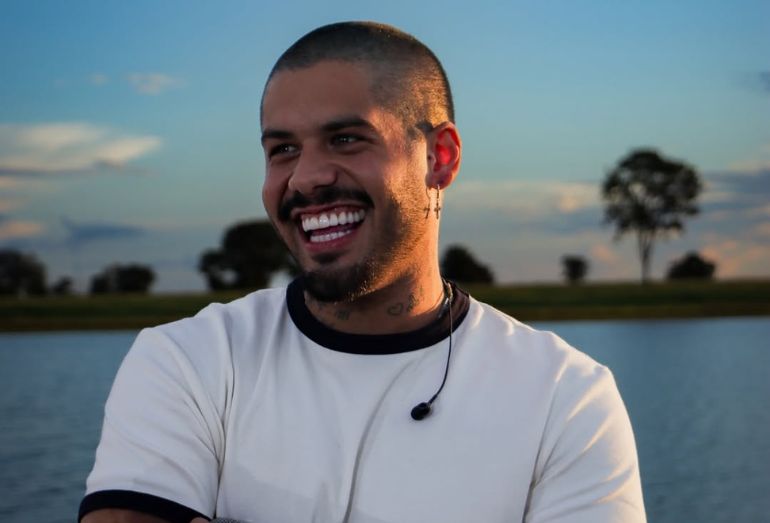 Um plano médio do cantor Zé Felipe, sorrindo abertamente e olhando para a direita. Ele está com a cabeça raspada, barba aparada e usa uma camiseta branca com golas e punhos pretos. Ele veste um brinco de argola com uma cruz pendurada na orelha esquerda e um fone de ouvido de retorno pendurado no pescoço. O fundo é um lago e vegetação desfocada sob o céu do pôr do sol.
